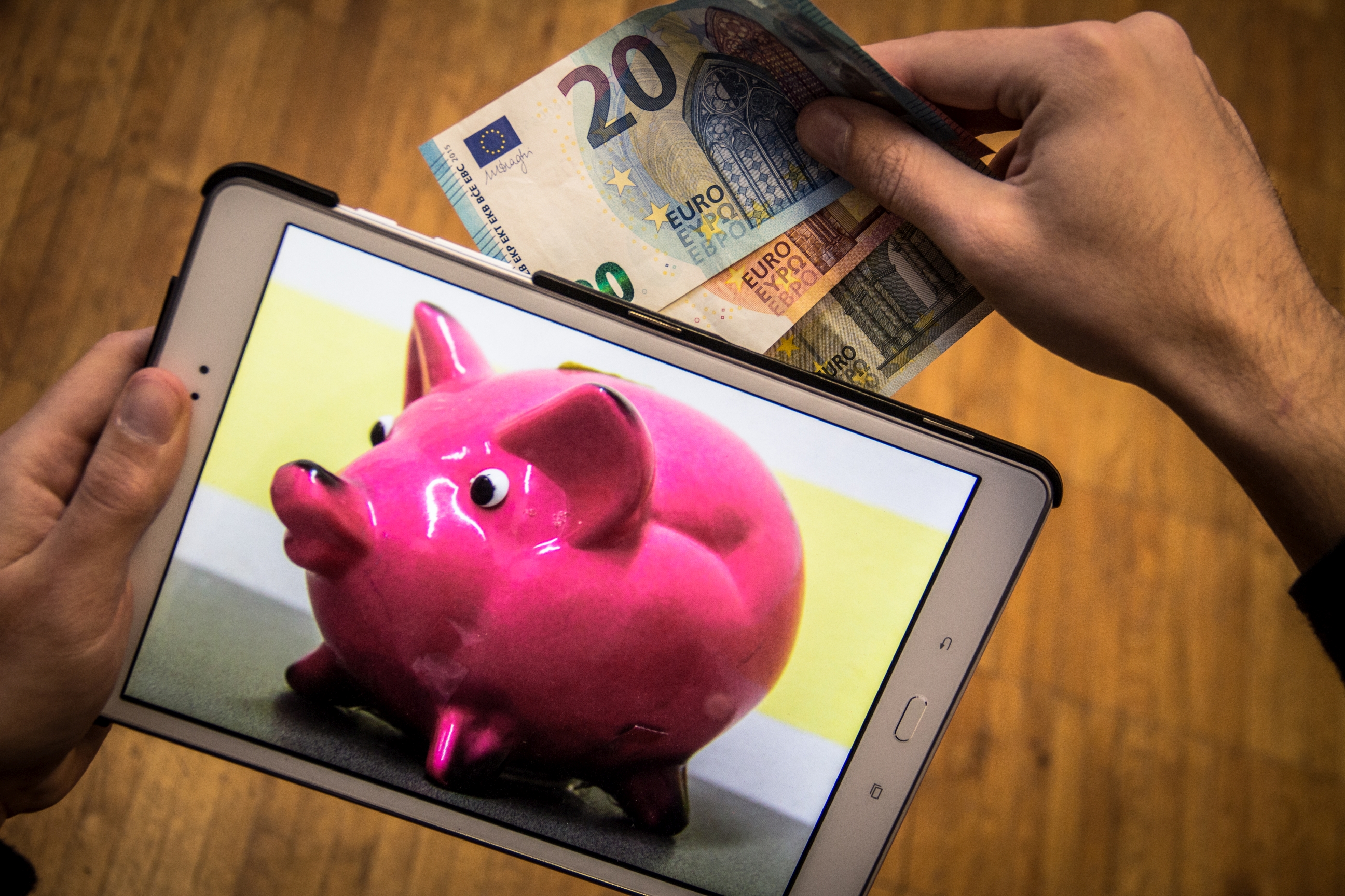 Auf einem Tablet ist ein Sparschwein abgebildet. Eine Hand steckt Geldscheine in das Sparschwein.