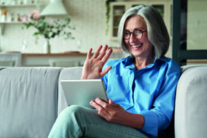 Happy 60s older mature middle aged adult woman waving hand holding digital tablet computer video conference calling by social distance virtual family online chat meeting sitting on couch at home.