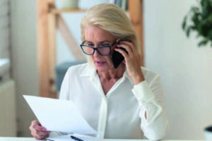 Concentrated senior businesswoman in glasses sit at office desk talk on cellphone reading paperwork document, focused middle-aged woman speak with client on smartphone discuss contract