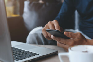 Eine Person sitzt mit dem Smartphone in der Hand vor einem aufgeklappten Laptop.