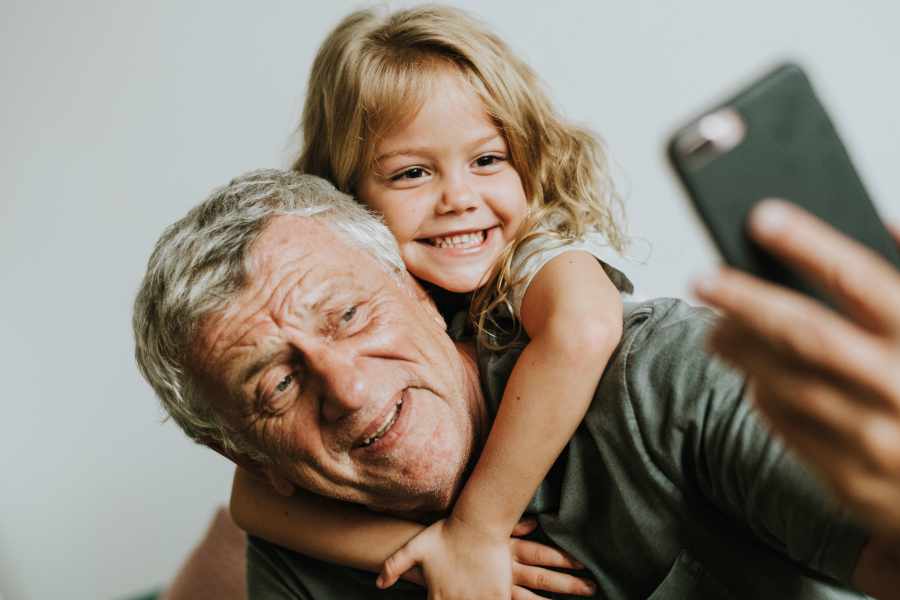 älterer Mann mit kleinem Mädchen auf dem Rücken lächeln beide in die Smartphone-Kamera