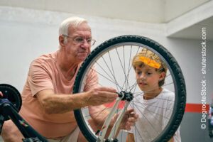 Attentive senior man in eyeglasses with cute grandson repairing wheel of modern bicycle while spending time together on parking lot