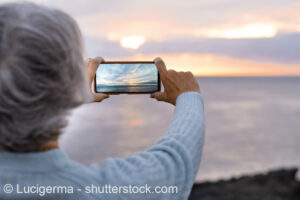 Eine Frau fotografiert mit ihrem Smartphone den Sonnenuntergang am Meer.