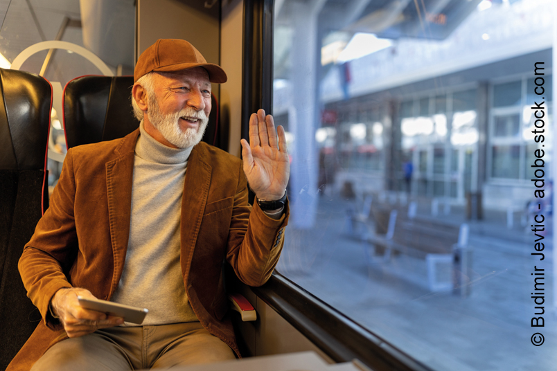 Ein älterer Mann sitzt mit seinem Smartphone in der Hand im Zug und winkt zum Abschied.