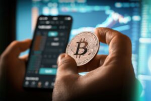 Eine Person hält eine Münze mit Bitcoin-Logo vor einen Bildschirm mit Börsenkursen.