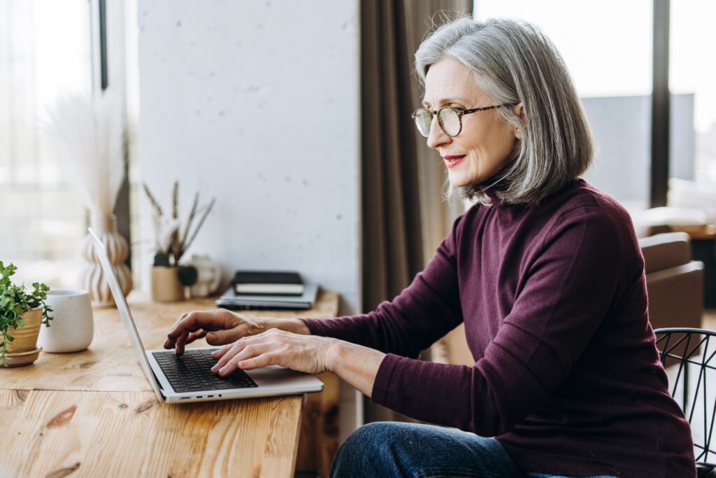 Interessiert schauende ältere Frau mit grauen Haaren sitzt vor einem Laptop und tippt