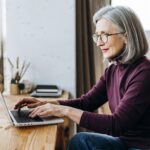 Interessiert schauende ältere Frau mit grauen Haaren sitzt vor einem Laptop und tippt