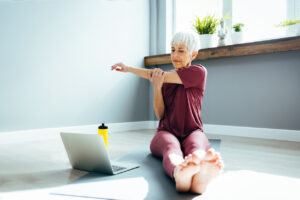 Beautiful senior woman doing stretching exercise while sitting on yoga mat at home.